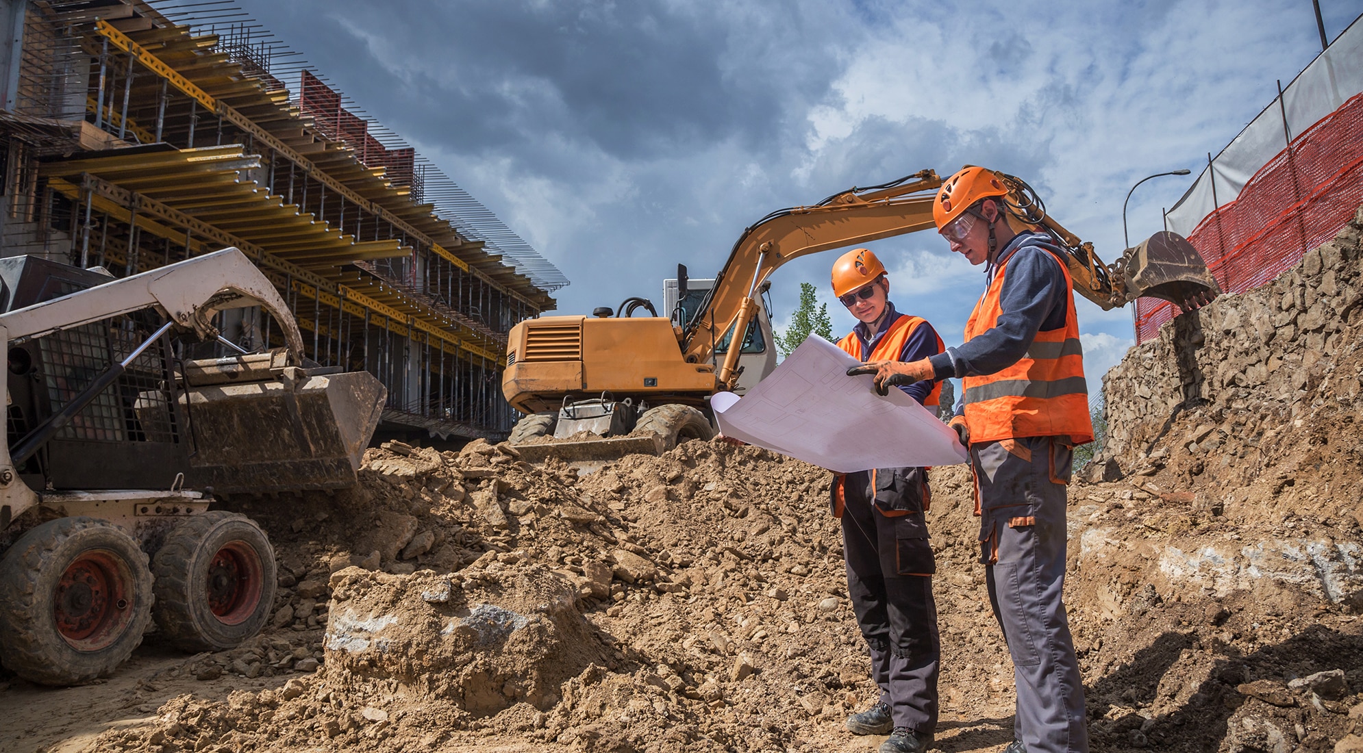 Two construction workers in hard hats stand in front of a construction site, with scaffolding and machinery in the background.